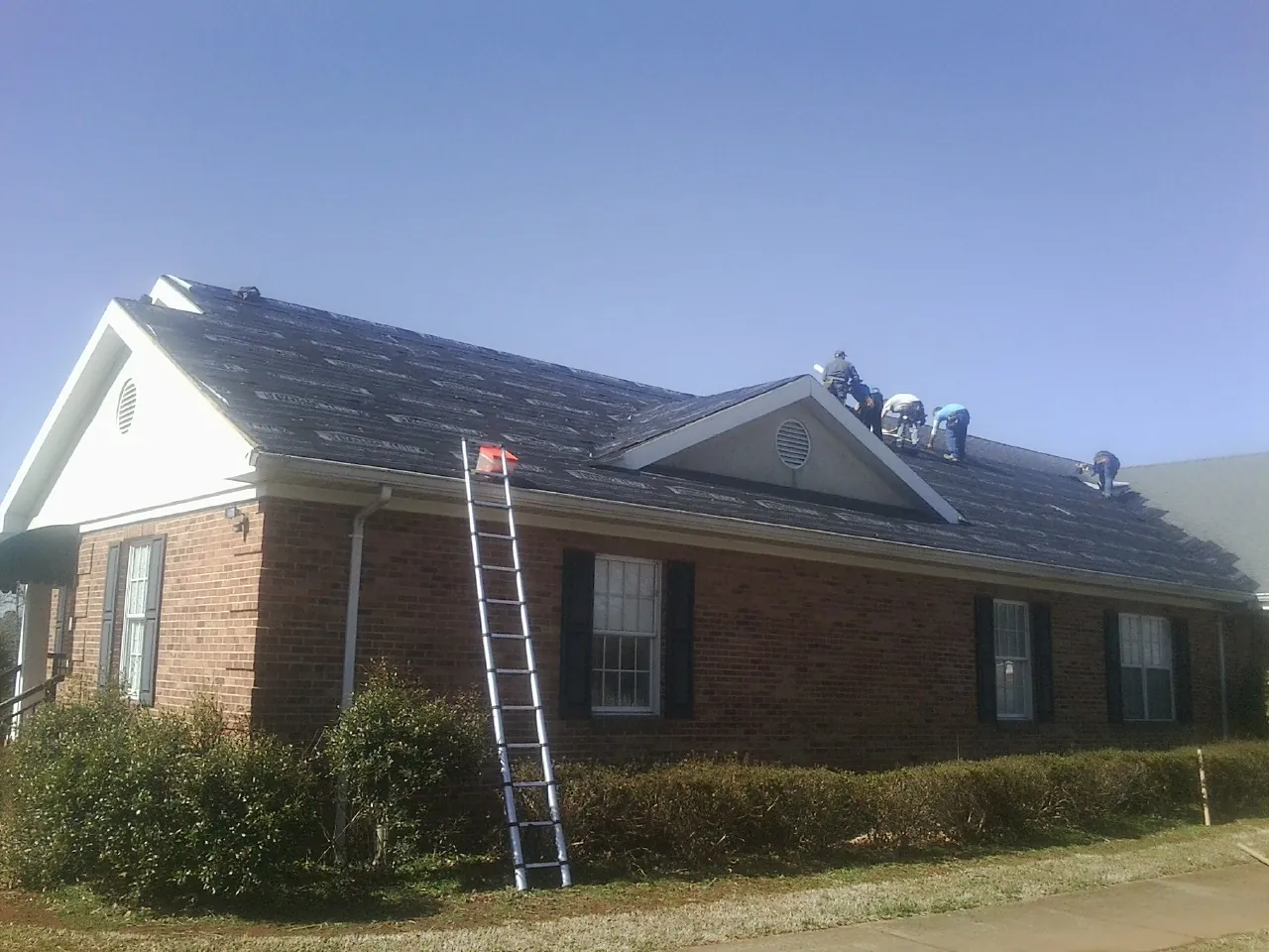 Crew tearing shingles off a brick ranch roof with a ladder set — Emergency Roof Repair work by our crew in Bejou