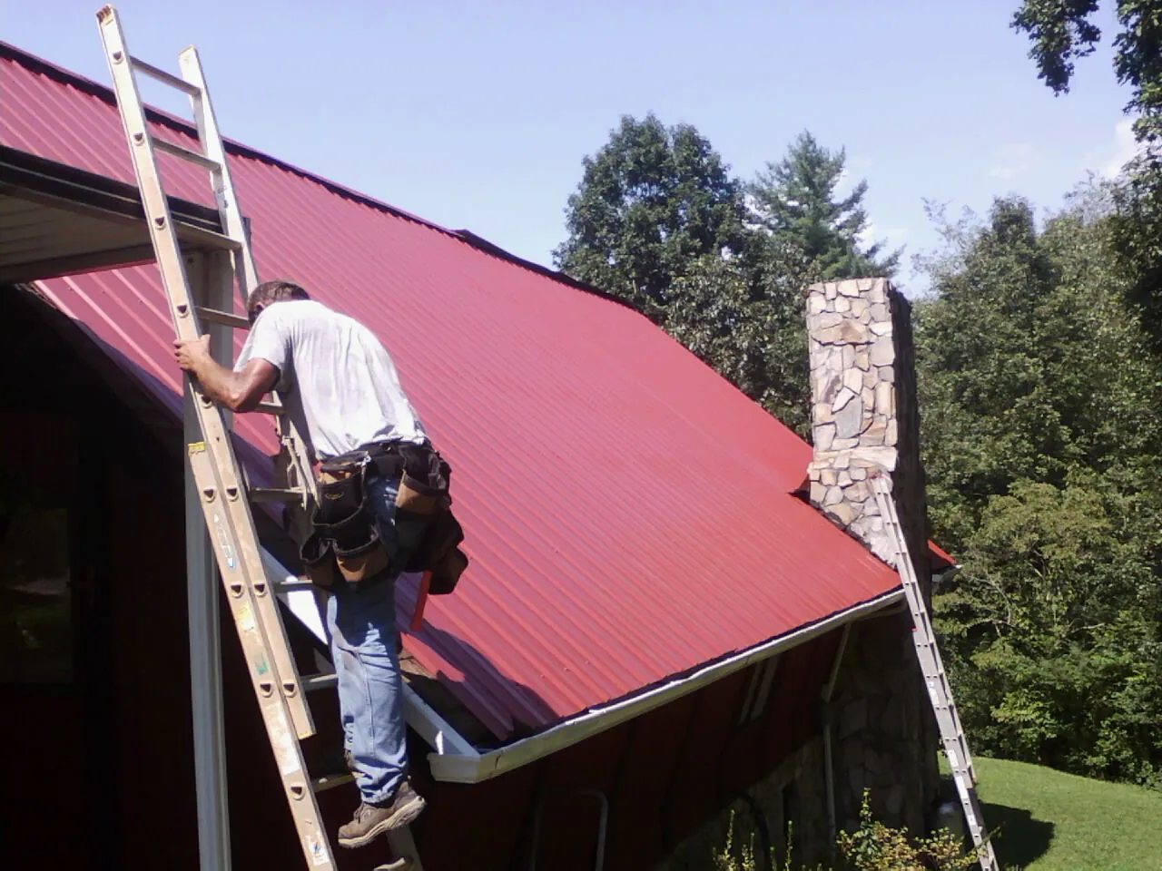 Roofer on a ladder beside a red metal roof and stone chimney — Soffit & Fascia Repair by Roofing Brothers in Bejou